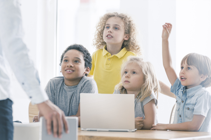 Auf der Abbildung sind Schülerinnen und Schüler zu sehen, die vor einem Laptop sitzen. Ein Junge hebt die Hand, weil er sich zu Wort melden möchte.