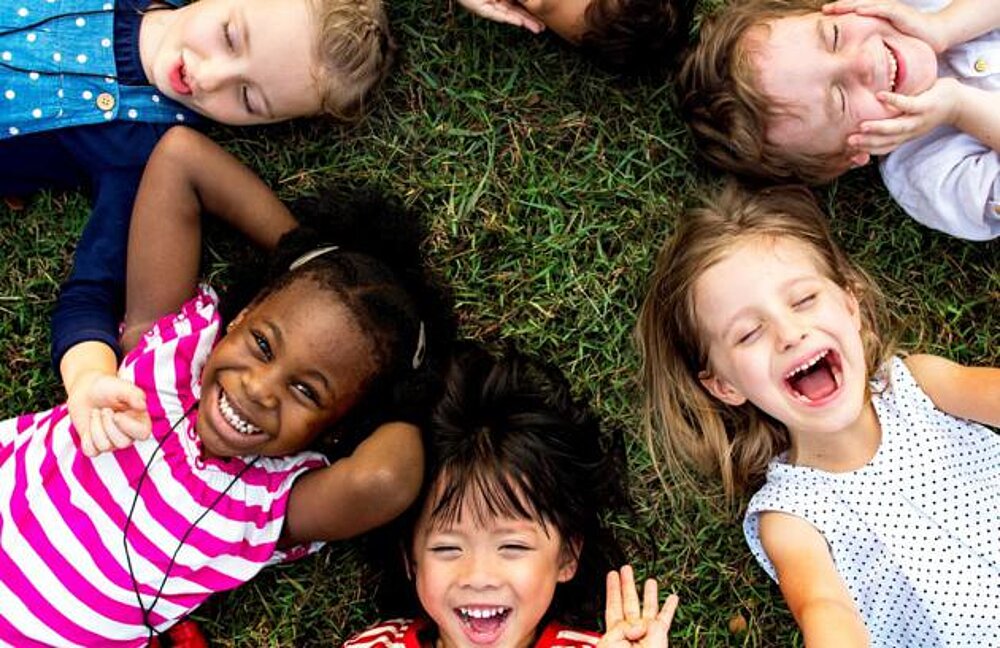 Abbildung: Kinder aus der Vogelperspektive, die zugewandt auf einer Wiese liegen und fröhlich in den Himmel sehen