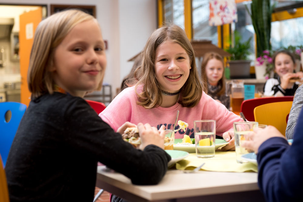 Abbildung zeigt Schülerinnen und Schüler, die in einer Schulmensa sitzen und zu Mittag essen.