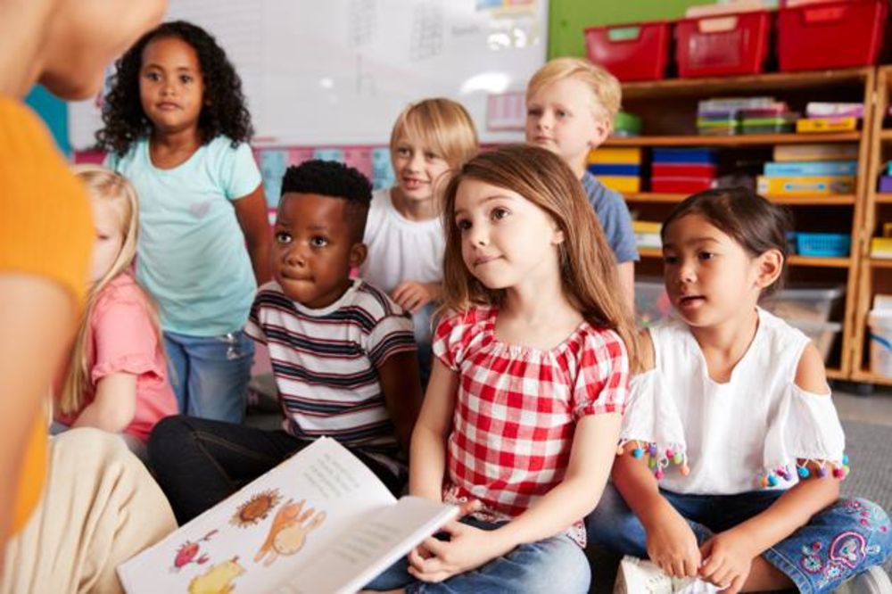 Abbildung zeigt Schülerinnen und Schüler in einem Raum sitzen. Sie hören einer Lehrerin zu, die Ihnen aus einem Buch vorliest.