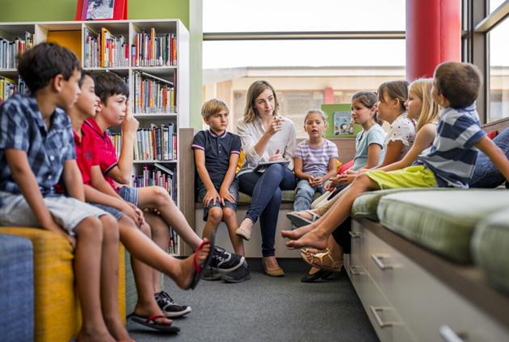 Abbildung zeigt Schulklasse mit Lehrerin in Schulbibliothek. Abbildung zur Auflockerung der Seite.