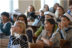 Abbildung zeigt Schülerinnen und Schüler zum Teil in historischer Kleidung, die in einem historisch nachempfundenen Klassenzimmer sitzen.